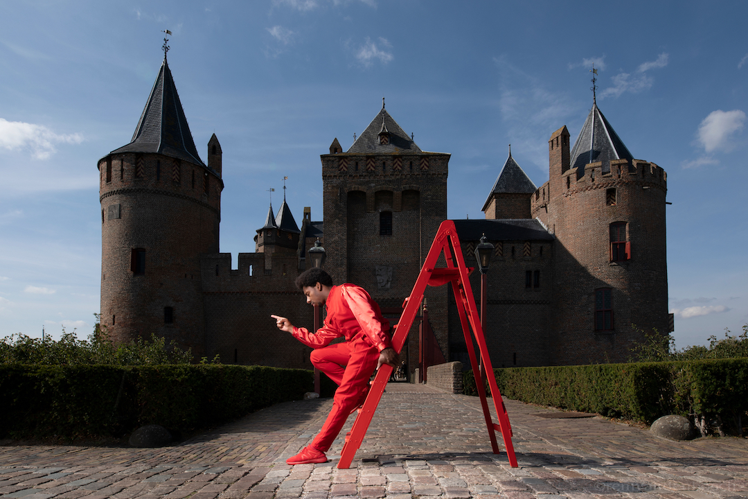 Man in het rood staat naast een trap in een dramatische pose tijdens de performance Here comes the Flood. Op de achtergrond het Muiderslot.