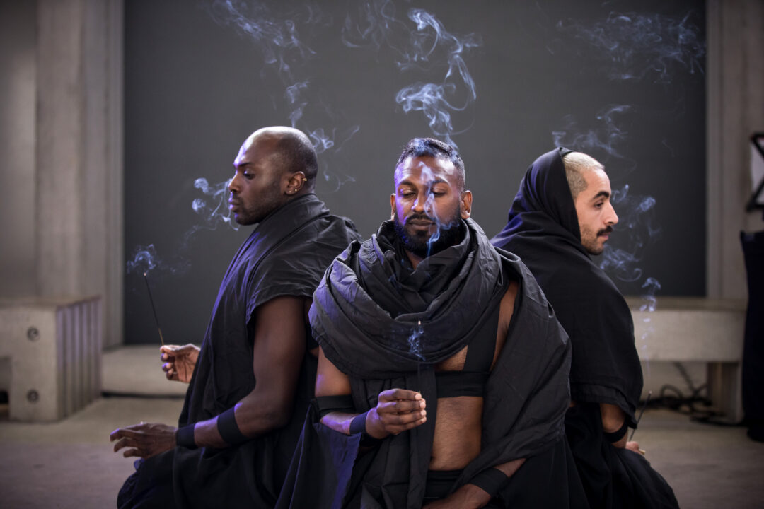 Three men sit back to back, with incense burning.