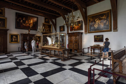 Gasten bezoeken de riddelzaal van het Muiderslot, met schilderijen aan de muur, kroonluchters en marmeren vloer.