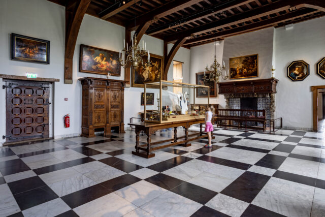 Een meisje bezoekt de riddelzaal van het Muiderslot, met schilderijen aan de muur, kroonluchters en marmeren vloer.