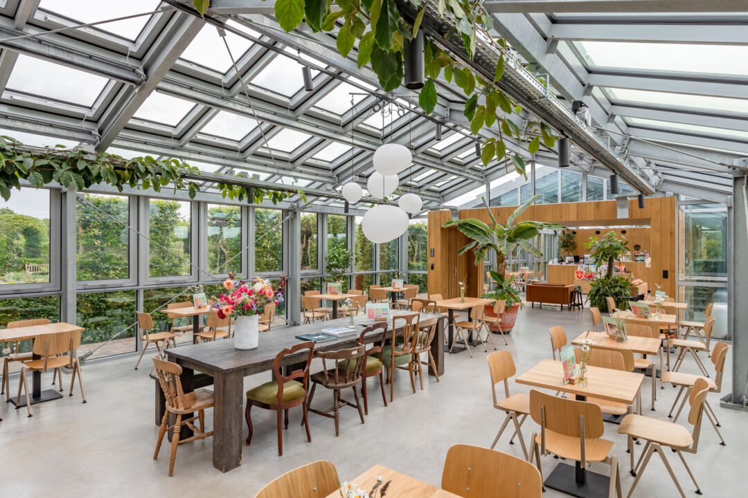 Het museumcafe de Tuinkamer heeft grote ramen en een glazen dak. De ruimte is ingericht met houten tafels en stoelen.
