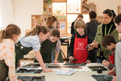 Kinderen zijn kunst aan het maken in het Kasteelatelier. Ze dragen schorten en staan in groepen om de tafels heen. Ze maken een linosnede, met zwarte verf.
