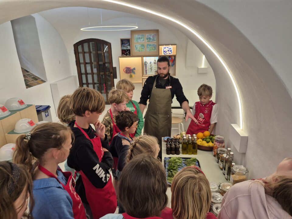 Een groep kinderen krijgt les van de kasteelkok. Ze krijgen uitleg over ingrediënten. Alle kinderen dragen een rode Muiderslot schort.