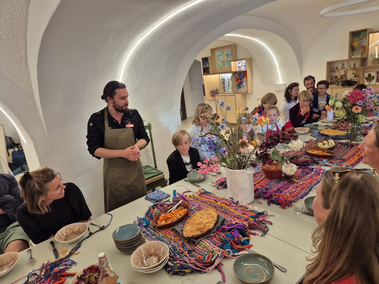 A group of children is about to start the Cooking Club, led by the castle chef. The table is set with colorful tableware.