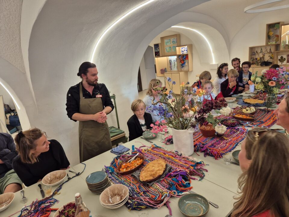 Een groep kinderen gaat beginnen aan de Kookclub, begeleid door de kasteelkok. De tafel is kleurrijk gedekt.