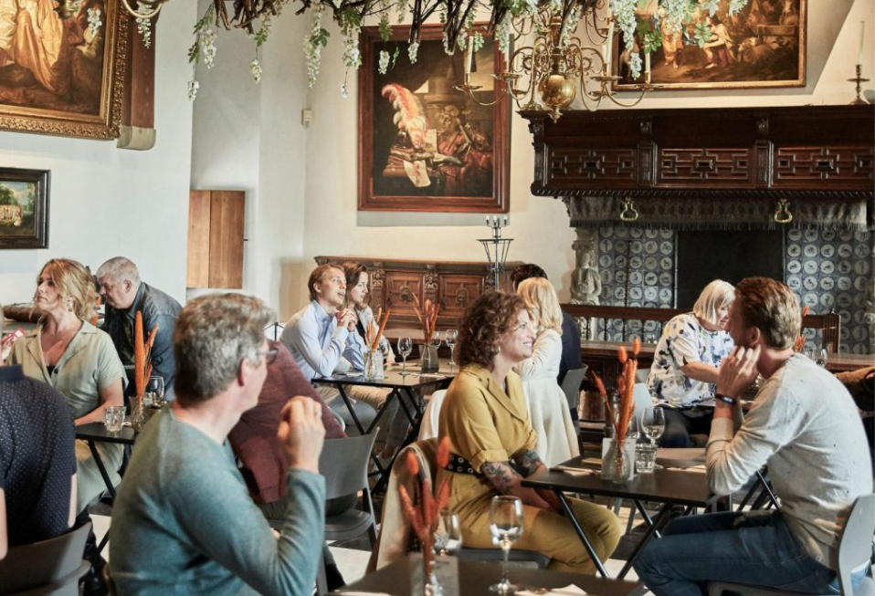 Tijdens het evenement 'de Maaltuin' zijn gasten aan het dineren. Er staan wijnglazen en bloemen op de tafels.