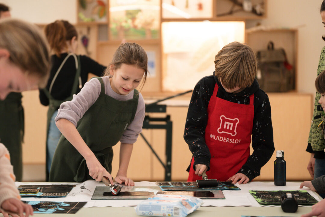 Kinderen zijn kunst aan het maken in het Kasteelatelier. Ze dragen schorten en staan in groepen om de tafels heen. Ze maken een linosnede, met zwarte verf.