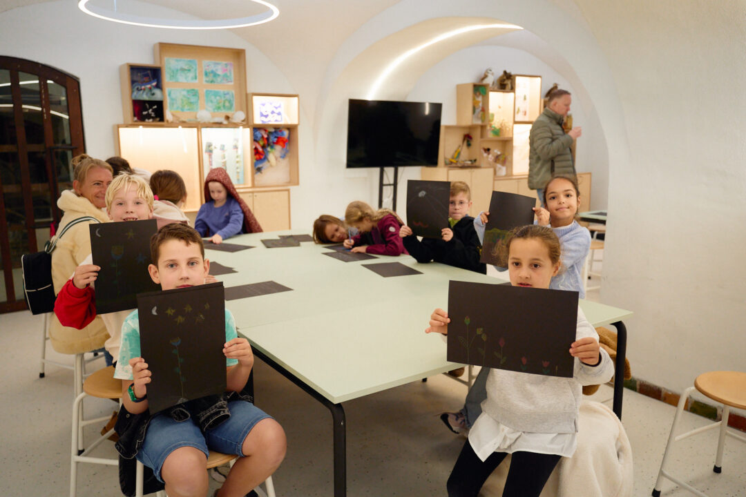 Een groep kinderen laat aan de camera hun kunstwerken zien. Het zijn tekeningen van bloemen op zwart papier.