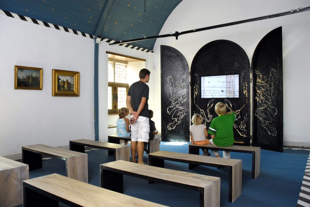 In het slotkapel zitten vier kinderen op een bankje een interactieve presentatie te bekijken. Het tv-scherm staat in een zwart drieluik met daarop witte tekeningen van leeuwen.