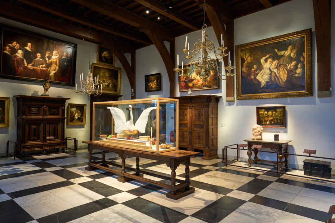 De riddelzaal van het Muiderslot, met schilderijen aan de muur, kroonluchters en zwart-witte marmeren vloer.