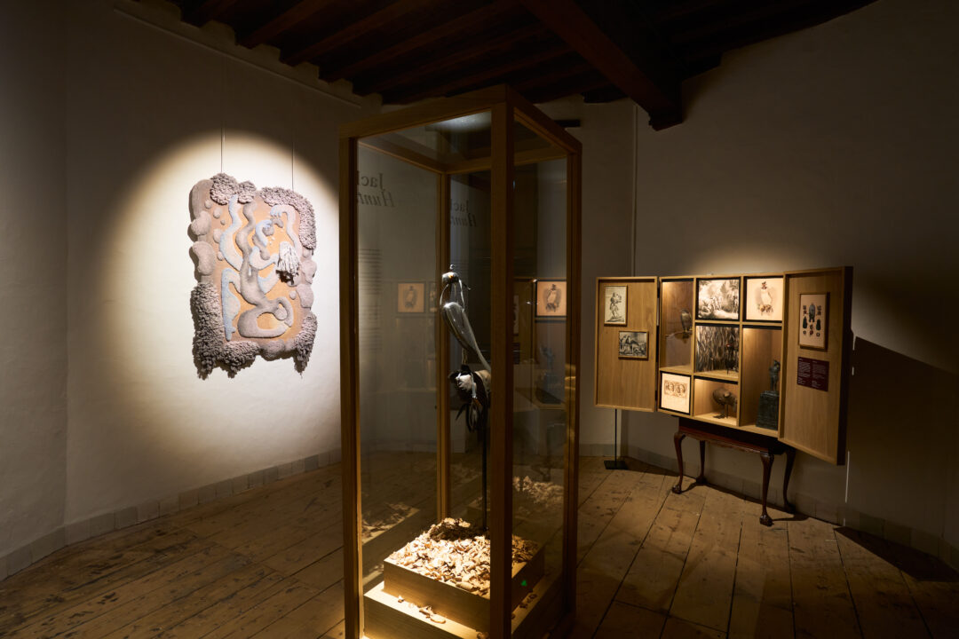 Tentoonstelling Soft!. Centraal in de kamer staat een glazen vitrine met een sculptuur. Aan de linkermuur hangt een wandkleed.
