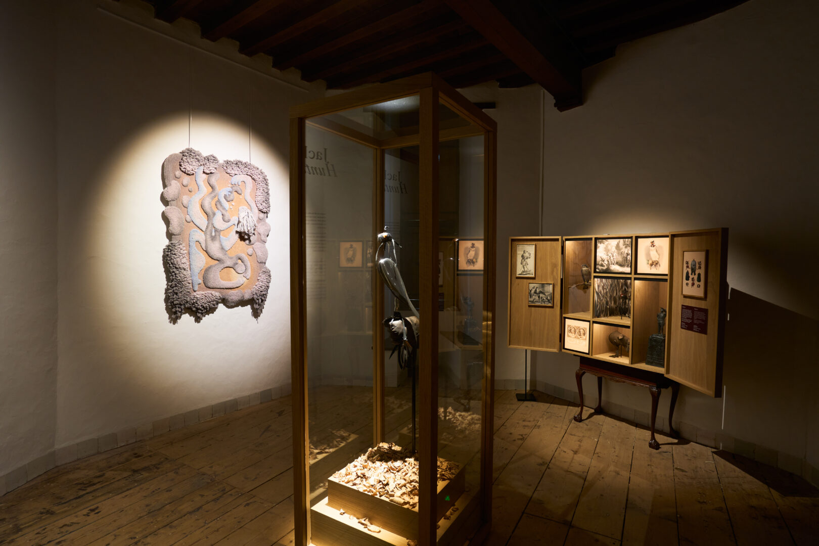 Tentoonstelling Soft!. Centraal in de kamer staat een glazen vitrine met een sculptuur. Aan de linkermuur hangt een wandkleed.