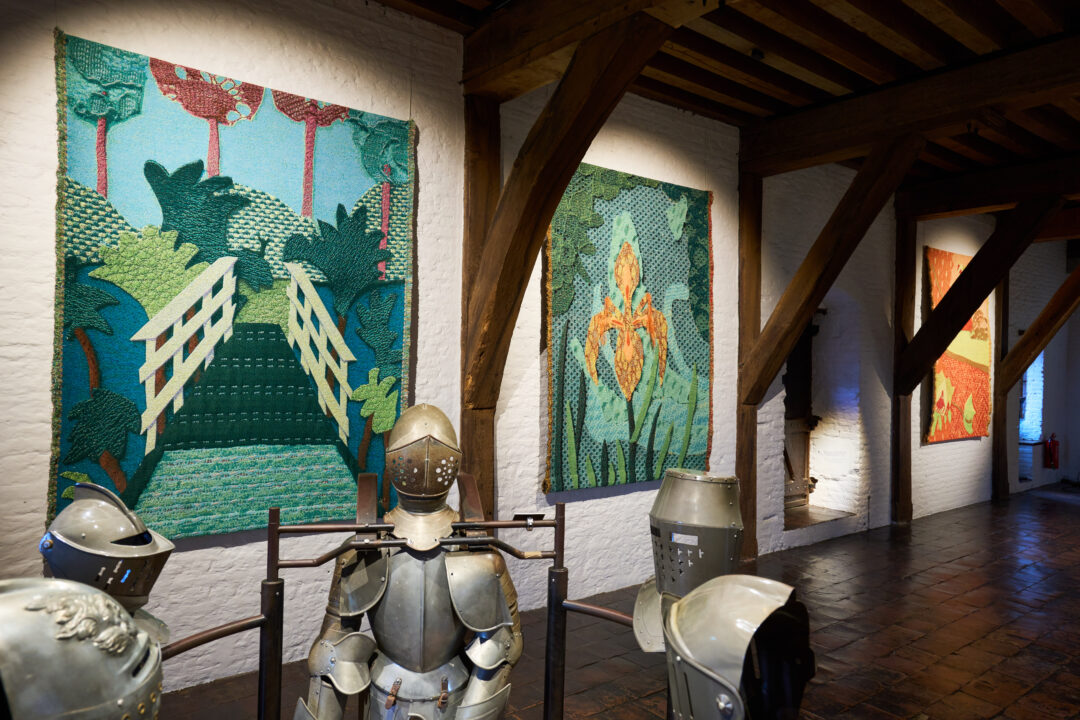 Tentoonstelling Soft!. Aan de wand hangen blauw-groene kleden van een bloem en een brug. In de zaal staan historische harnassen.