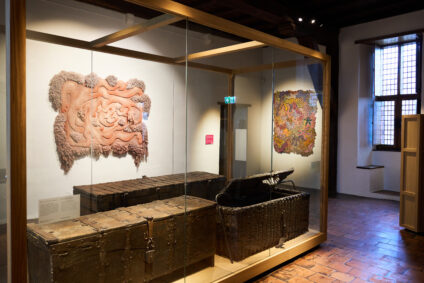 Tentoostelling Soft!. Aan de wand hangen grote, gestructureerde kleden. In het midden van de ruimte staat een glazen vitrine met grote donkerbruine kisten.