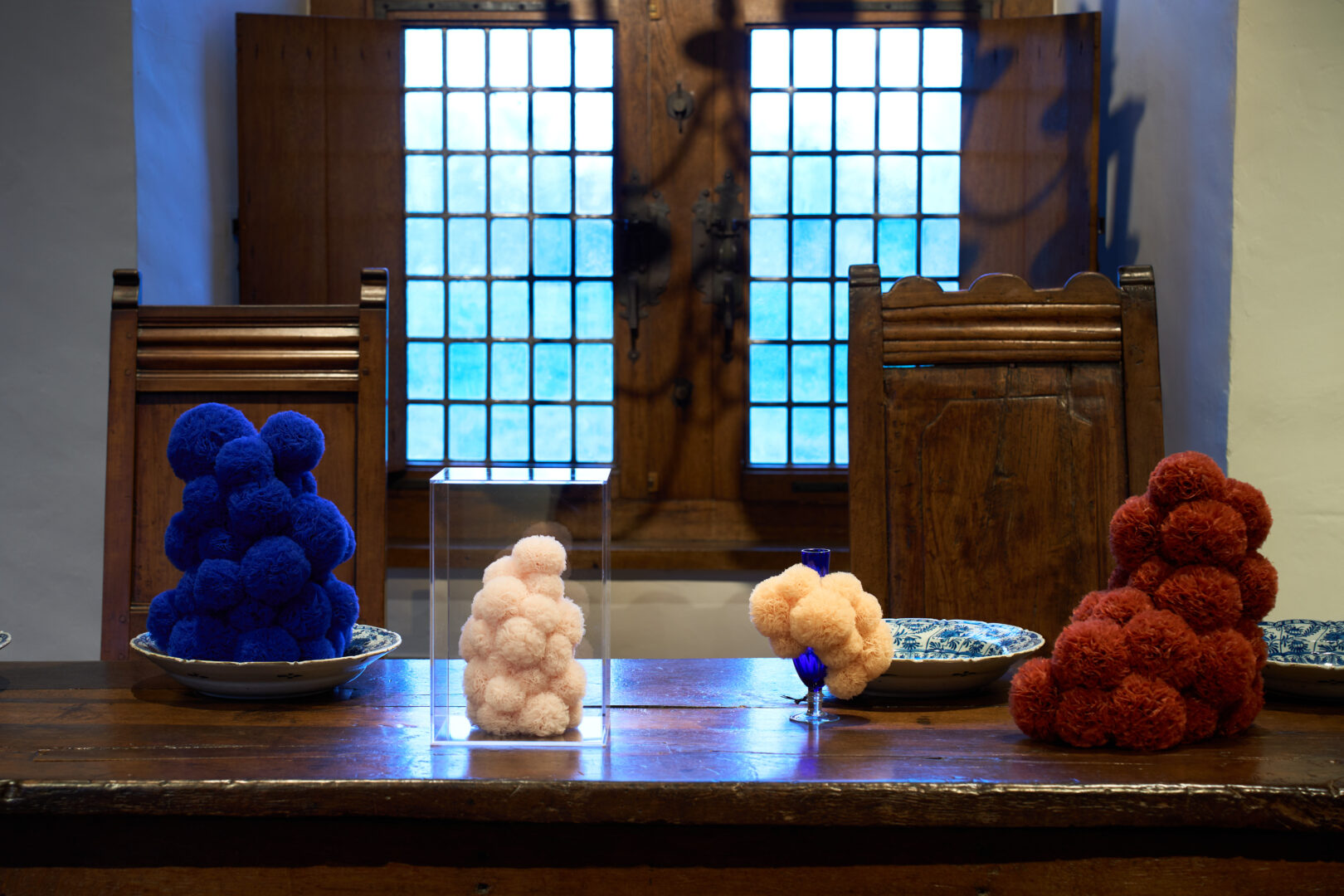 Een kunstwerk van de tentoostelling Soft!. Het zijn vier textielsculpturen die op een houten tafel staan, in blauw, roze, licht oranje en rood.