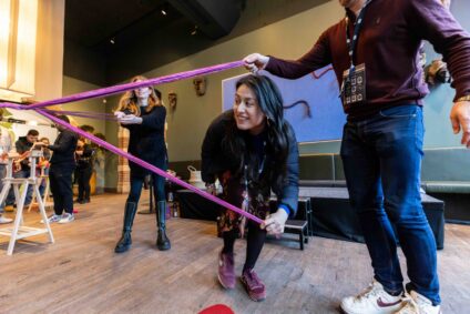 Een vrouw neemt deel aan de workshop 'In wol verweven'. Ze bukt en helpt met de creatie van een groot collectief textielkunstwerk. Het kunstwerk is paars.