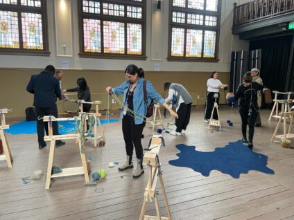 Mensen nemen deel aan de workshop 'In wol verweven'. De ruimte staat vol met wol en houten materialen om een groot interactief textielkunstwerk te maken.