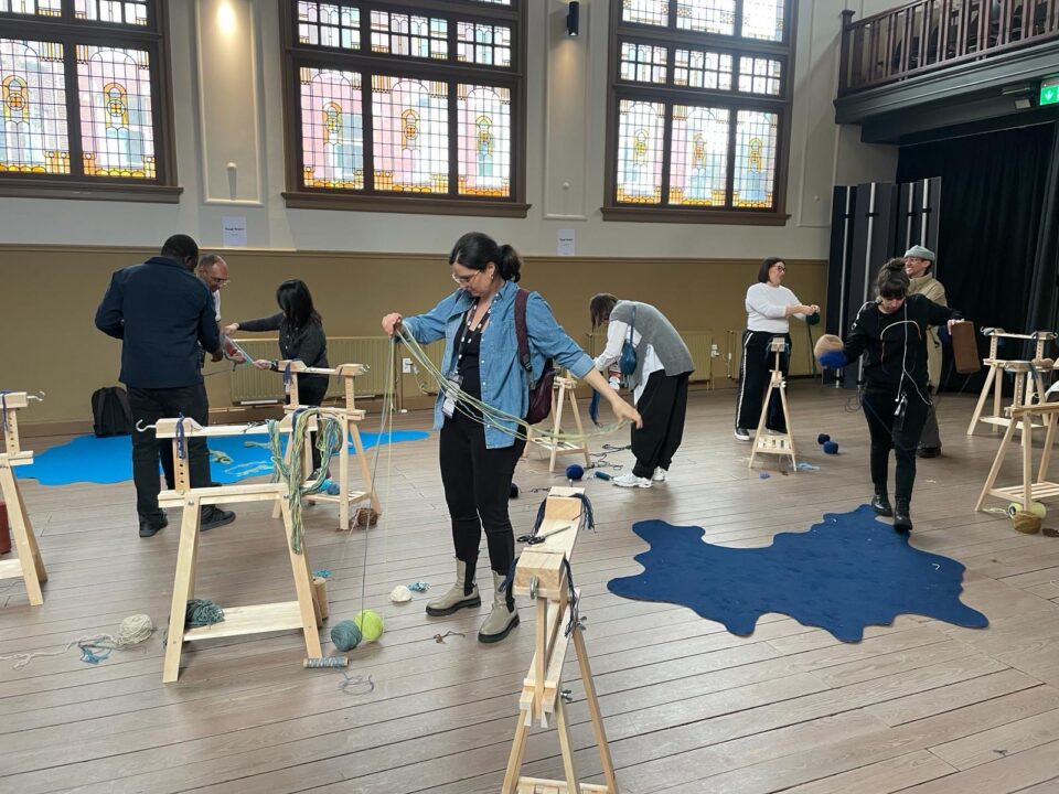 Mensen nemen deel aan de workshop 'In wol verweven'. De ruimte staat vol met wol en houten materialen om een groot interactief textielkunstwerk te maken.