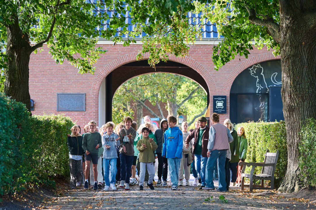 Een groep kinderen doet mee aan het educatieve programma Water: Vriend of Vijand? Ze luisteren naar de uitleg van Sefania Vitalis en lopen buiten. De zon schijnt.