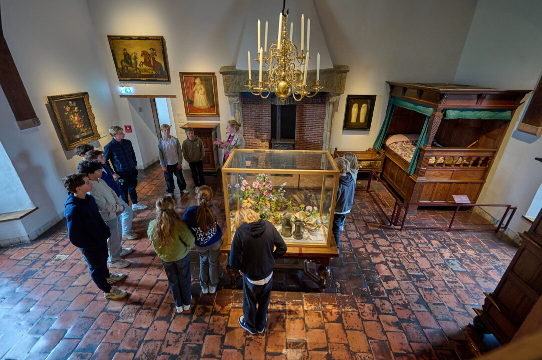 Een groep kinderen wordt rondgeleid in het Muiderslot door de museumdocent. Er hangt een grote kroonluchter in de ruimte.