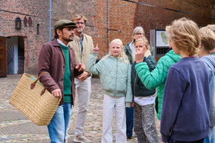 Een groep kinderen doet mee aan het educatieve programma Water: Vriend of Vijand? Ze luisteren naar de uitleg van Sefania Vitalis en spelen een spel. Sefania draagt een grote rieten mand.