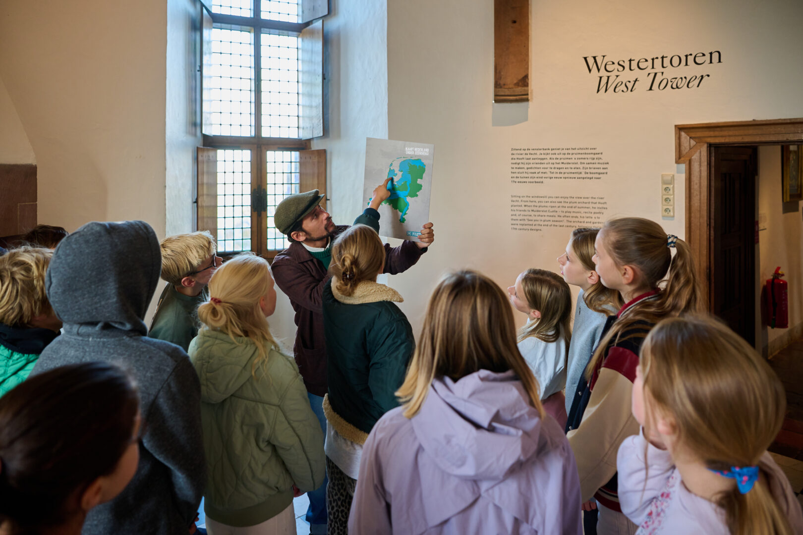 Een groep kinderen doet mee aan het educatieve programma Water: Vriend of Vijand? Ze luisteren naar de uitleg van Sefania Vitalis die een landkaart van Nederland vasthoudt.