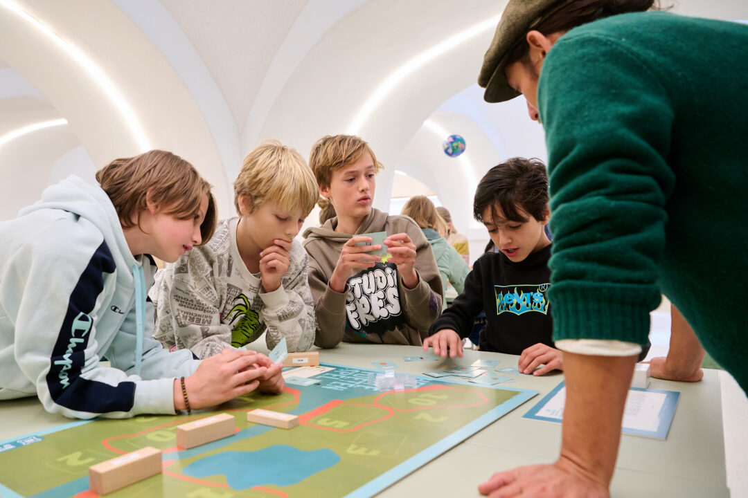 Een groep kinderen doet mee aan het educatieve programma Water: Vriend of Vijand? Ze staan rond een tafel en spelen een interactief bordspel.