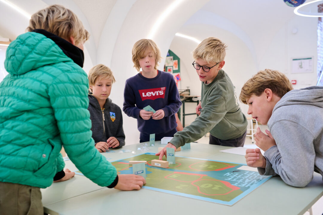 Een groep kinderen doet mee aan het educatieve programma Water: Vriend of Vijand? Ze staan rond een tafel en spelen een interactief bordspel.