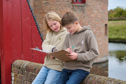 Twee kinderen doen mee aan het programma Oorlog en Vrijheid. Ze staan op de brug van het Muiderslot en houden klemborden vast om opdrachten te maken.