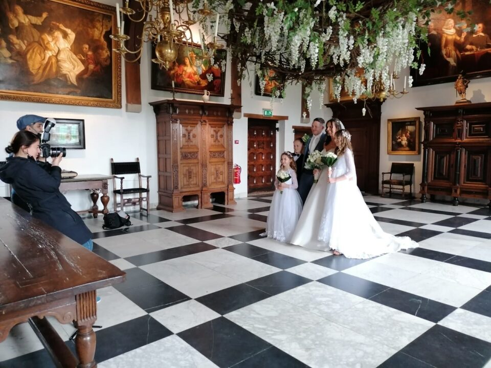 Een bruiloft in de Ridderzaal van het Muiderslot. Het bruidspaar, met drie kinderen, wordt gefotografeerd. Aan de kroonluchter hangen trossen van de witte regen.