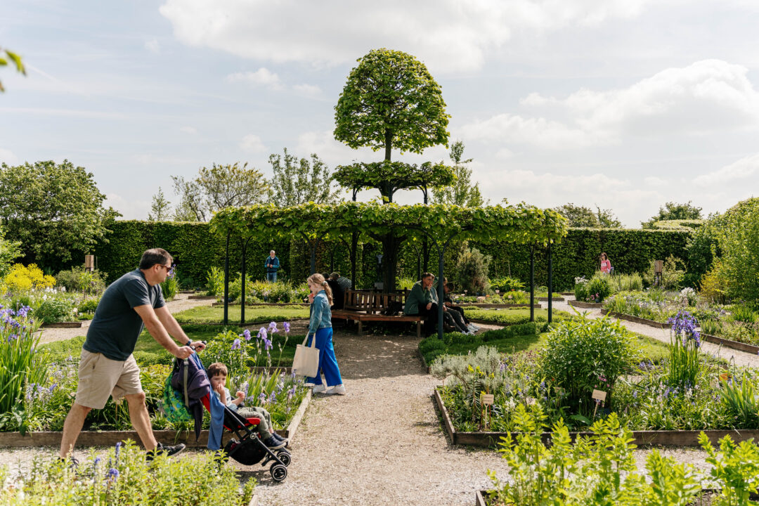 Gasten lopen rond in de tuin van het Muiderslot. De zon schijnt. Wat mensen zitten op het bankje in het pireel.
