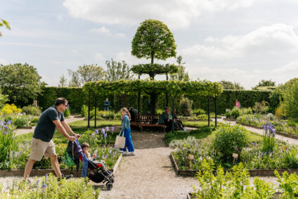 Gasten lopen rond in de tuin van het Muiderslot. De zon schijnt. Wat mensen zitten op het bankje in het pireel.