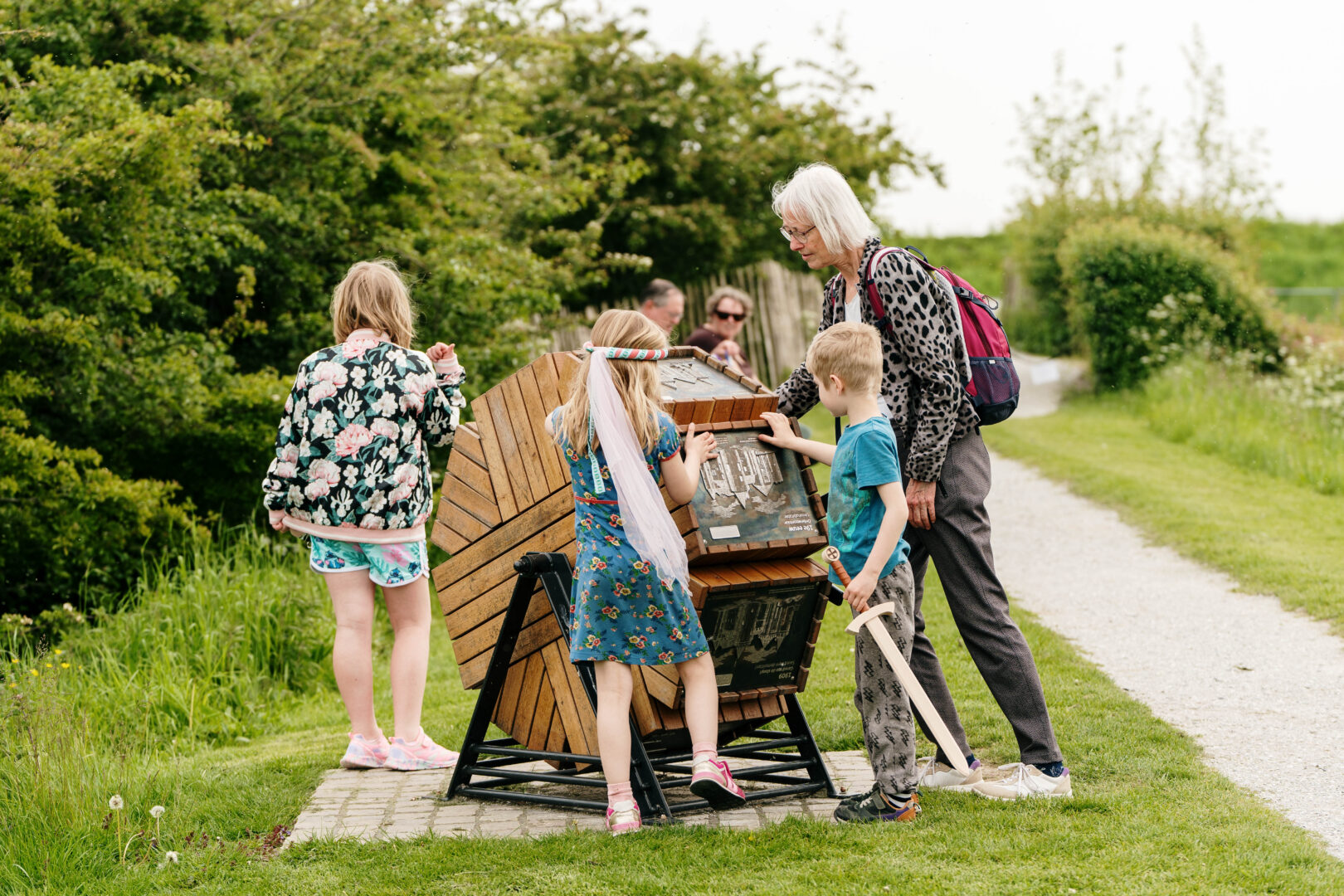 Kinderen zijn buiten aan het spelen met een groot, zeshoekig houten wiel dat informatieborden bevat. Een kindje houdt een speelgoedzwaard vast.
