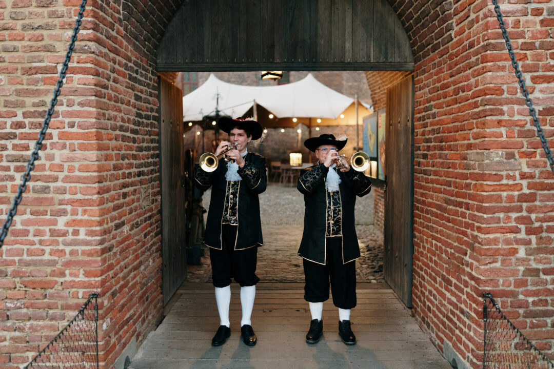 Twee als middeleeuwse heraut gekleedde mannen staan met trompet ontvangen gasten bij de poort van het kasteel.